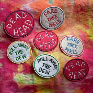 May include: A collection of embroidered patches with various phrases. The patches are circular and feature text like "DEAD HEAD", "FARE THEE WELL", and "ROLL AWAY THE DEW". The patches have different color combinations, including red, white, and green.