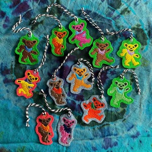 May include: A selection of colourful bear-shaped felt ornaments with white and black string hangers. Each bear is a different colour, including brown, yellow, pink, and orange, set against a green or denim-coloured background.