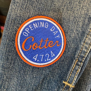 May include: A blue and orange circular patch sewn onto denim fabric. The patch says "Opening Day Cotter 4.7.24" in white text.