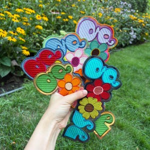 May include: A collection of embroidered patches featuring the word "one" in a stylized, rounded font layered over various colorful flower shapes. The patches are rendered in vibrant shades of red, blue, green, orange, yellow, and purple.