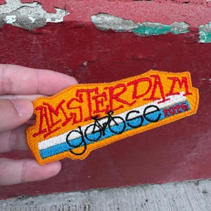 May include: Embroidered patch with the word "AMSTERDAM" in red and orange, with a bicycle graphic and the word "goose" in blue. The patch has a white and blue stripe design.