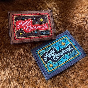 May include: Two embroidered patches with the text "Merry Goosemas 2025". One patch is brown with red and gold accents, the other is blue with turquoise and gold accents. Both have a rectangular shape.
