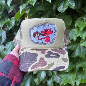 May include: A camouflage-brimmed trucker hat with a tan crown. The hat has an embroidered patch of a red devil holding a bottle, set against a blue cloud. The hat is held by a person.