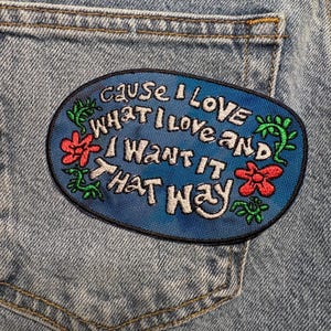 May include: An embroidered patch with the text "Cause I love what I love and I want it that way" in white on a blue oval. The patch is decorated with red flowers and green vines, set against a denim background.