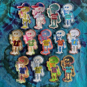 May include: A collection of embroidered patches featuring skeleton figures in various colors and outfits. Some skeletons wear baseball caps, t-shirts with text like "GOOSE 4EVER" and "DEAD 4EVER", and are giving the peace sign. The patches are on a tie-dye background.