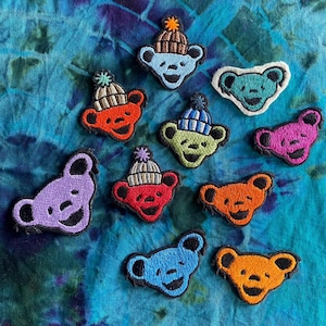 May include: A collection of embroidered bear patches in various colors, including blue, orange, red, green, and purple. Some bears wear knit hats with pom-poms. The patches are arranged on a tie-dye blue and green fabric.