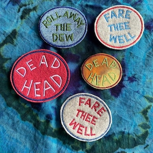 May include: Five embroidered patches with text on a tie-dye background. The patches are oval and circular, with phrases like "Roll Away The Dew" and "Fare Thee Well" in various colors. The patches are likely for clothing or accessories.