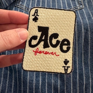 May include: Embroidered patch in the shape of an Ace playing card. The card has a beige background with black text that reads "Ace" and red text that reads "forever". The patch also features the spade and club symbols.