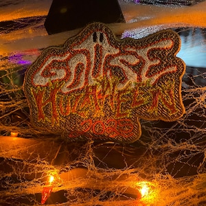 May include: Embroidered patch in the shape of a ghost with the words "Ghost Halloween 2023" in a spooky font. The patch is primarily brown with red, yellow, and white accents. The background includes cobwebs and orange lights.