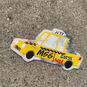 May include: Embroidered patch in the shape of a yellow New York City taxi cab. The patch features the text "NYC", "MSG", "GOOSE", and "2024". The taxi has black wheels and a white border.