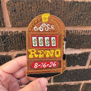 May include: An embroidered patch shaped like a slot machine with a brown frame, a yellow top accent, and white reels displaying green dollar signs. The text RENO is in yellow, and the date 8-16-26 is in white on a red background.