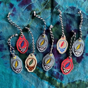 May include: A collection of embroidered tags featuring a skull with a lightning bolt and a colorful design. The tags are made of denim and various colors, with a black and white twisted cord for hanging. The tags are on a blue tie-dye background.