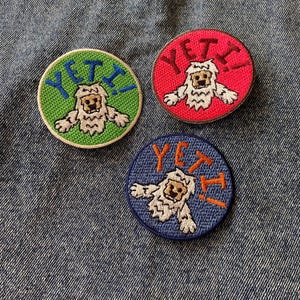 May include: Three round embroidered patches featuring a cartoon yeti design. One patch is green with blue text, another is red with red text, and the third is blue with orange text. The word "YETI" is displayed on each patch.