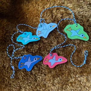 May include: Five colorful felt bird ornaments with blue, green, pink, and teal backgrounds. Each bird has a light blue body, orange beak, and yellow feet. The ornaments are connected by a blue and white twisted cord.