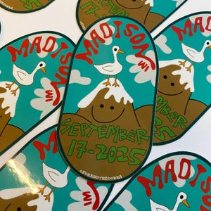 May include: Oval sticker with a teal background featuring a mountain with a smiling face and a white duck on top. The sticker reads "MADISON WI" in red and "SEPTEMBER 17-2025" in green. The sticker has a brown border.