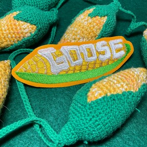 May include: Close-up of handmade crochet corn on the cob ornaments with green husks and yellow kernels. An embroidered patch with the word "GOOSE" in white letters is also visible. The ornaments are on a green background.