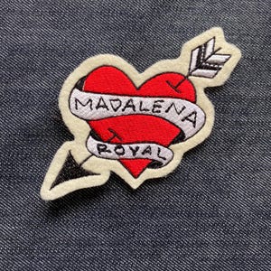 May include: A heart-shaped patch with a red center, pierced by an arrow. The patch has a white banner with the words "MADALENA ROYAL" in black lettering. The patch is outlined in white and set against a denim background.