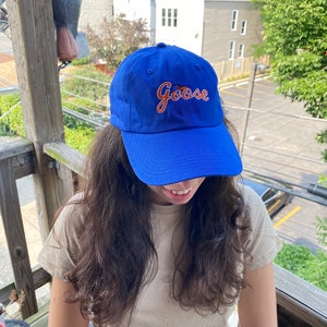 May include: A royal blue baseball cap featuring the word Goose embroidered in orange cursive script with a white outline on the front panel. The cap has a curved brim and a structured crown with ventilation eyelets at the top of the fabric panels.