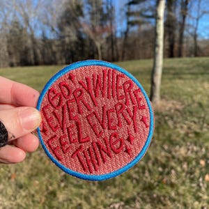 May include: A round, embroidered patch with the words "GO EVERYWHERE FEEL EVERYTHING" in red script. The patch has a red background and a blue border. The patch is held up in front of a blurred outdoor background.