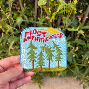 May include: A square embroidered patch with rounded corners featuring a light blue sky background, a yellow sun, and four green pine trees. The red text reads FROST AMPHITHEATER, with the white text GOOSE vertically on the left and 26 on the right.