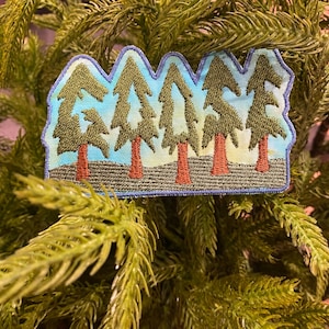 May include: Embroidered patch with the word "FORREST" spelled out using five green pine trees with brown trunks against a blue sky background.