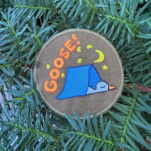 May include: A circular embroidered patch with a brown background featuring a blue tent-like shape covering a white goose head with an orange beak. The text "GOOSE!" is stitched in orange above the tent, accompanied by small yellow stars and a crescent moon.