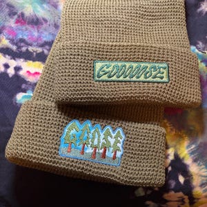 May include: Two olive green knit beanies with embroidered patches. One patch reads "GOODOOSE" in green lettering. The other patch features a forest scene with trees and the word "GOOSE" in a stylized font.