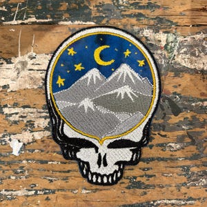 May include: Embroidered patch featuring a skull design with a mountain and night sky scene. The patch has a blue background with a yellow crescent moon and stars, and gray mountains. The skull is black and white with a yellow border.
