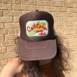 May include: A brown trucker hat with a mesh back and a white rectangular patch on the front. The patch features an embroidered yellow corn cob with green husks and the red cursive text Cotter's corn club.