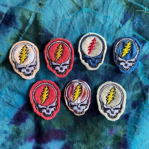 May include: Eight embroidered patches with a skull and lightning bolt design. The patches are in various colours, including red, blue, and cream, with yellow and red lightning bolts. The patches are displayed on a tie-dye background.
