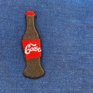 May include: Embroidered patch in the shape of a soda bottle with a red label and the word "Goose" in white script. The bottle is brown with a red cap and is set against a blue denim background.