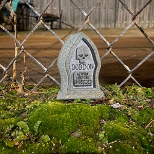 May include: A gray tombstone with a skull and crossbones design and the text "Bob Don He Who Lies Here Undecided". The tombstone is sitting on a bed of green moss.