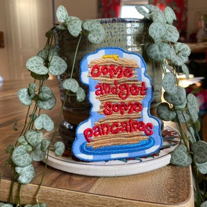 May include: A blue and white checkered patch with a stack of pancakes embroidered on it. The pancakes are brown and have a yellow dollop of butter on top. The text "Come and get some pancakes" is embroidered in red on the pancakes.