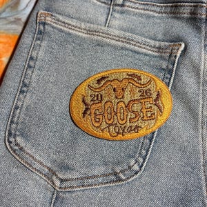 May include: An embroidered patch with the text "GOOSE TEXAS" in a gold and brown color scheme. The patch is oval-shaped and features a bull's head and the numbers "21" and "26". The patch is sewn onto a light blue denim pocket.