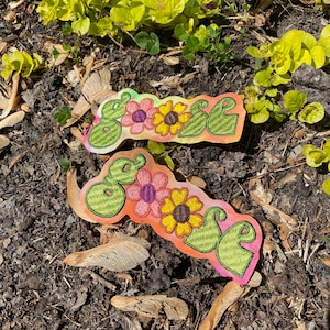 May include: Two embroidered patches with the word "SOUL" in green, with pink, yellow, and purple flowers. The patches are on a colorful background of orange, pink, and green. The patches are lying on the ground.