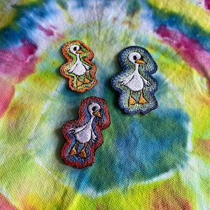 May include: Three embroidered iron-on patches of geese in different colors. The geese are white with orange beaks and feet. The patches are outlined in orange, blue, and red.