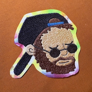 May include: Embroidered patch featuring a stylized face with a beard, sunglasses, and a backward baseball cap. The patch has a tie-dye border with a black cap and brown beard. The patch is on a brown surface.