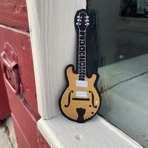 May include: A gold and black embroidered patch of a hollow-body electric guitar. The guitar has a black pickguard and white knobs.