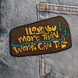 May include: Embroidered patch with the text "I Love You More Than Words Can Tell" in gold lettering on a brown background. A blue river and green trees are also embroidered on the patch. The patch is sewn onto a denim fabric.