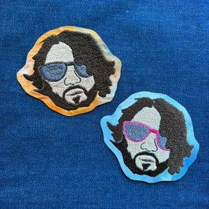 May include: Two embroidered patches featuring a bearded man with sunglasses. One patch has a yellow, orange, and brown border, while the other has a light blue border with pink-rimmed sunglasses. The patches are on a blue denim background.