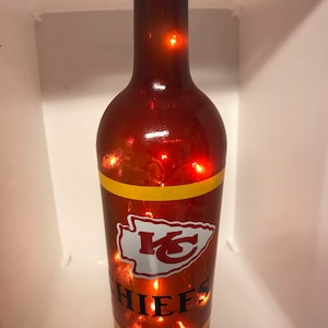May include: A brown glass wine bottle with a red and yellow Kansas City Chiefs logo and the word "CHIEFS" on it. The bottle is decorated with string lights.