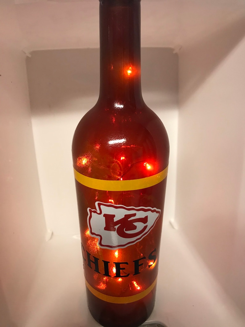 May include: A brown glass wine bottle with a red and yellow Kansas City Chiefs logo and the word "CHIEFS" on it. The bottle is decorated with string lights.