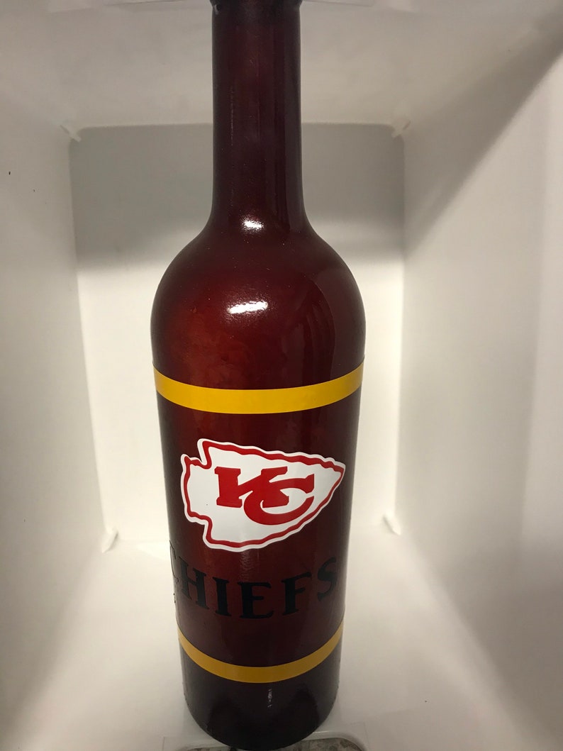 May include: A brown glass wine bottle with a red, white, and gold Kansas City Chiefs logo and the word "CHIEFS" in black lettering.
