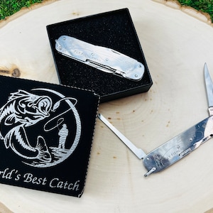 May include: Two silver multi-tool pocket knives with the words "Fly Fisherman" engraved on them. One knife is in a black box with a black leatherette case with a silver fish and fisherman design and the words "World's Best Catch".