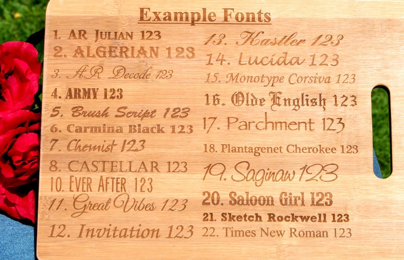 May include: A light-colored wooden cutting board with a handle, showcasing various font examples. The text "Example Fonts" is at the top, followed by numbered font styles like "AR JULIAN 123" and "ALGERIAN 123". The board is surrounded by a blurred green and red background.