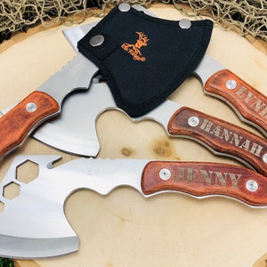 May include: A collection of hatchets with wooden handles and stainless steel blades, some engraved with names. One hatchet has a bottle opener and a black sheath with the Elk Ridge logo. The hatchets are arranged on a wooden surface.
