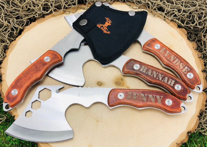 May include: A collection of hatchets with wooden handles and stainless steel blades, some engraved with names. One hatchet has a bottle opener and a black sheath with the Elk Ridge logo. The hatchets are arranged on a wooden surface.