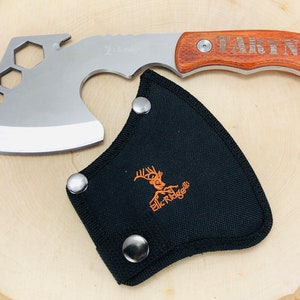 May include: A silver and wood hatchet with a black sheath. The hatchet has a wooden handle engraved with the name "TARYN". The sheath is black with the "Elk Ridge" logo in orange.