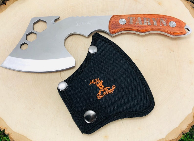 May include: A silver and wood hatchet with a black sheath. The hatchet has a wooden handle engraved with the name "TARYN". The sheath is black with the "Elk Ridge" logo in orange.