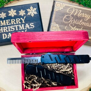 May include: A black comb and knife set inside a red wooden box, presented on a wooden surface. Two additional wooden boxes with engraved text and snowflake designs are visible. The text on the boxes reads "Merry Christmas Dad" and "Merry Christmas Stephen".